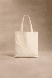 Un sac fourre-tout en toile beige unie avec deux poignées se tient debout sur un fond neutre et texturé.