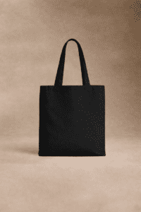 Un sac fourre-tout noir simple avec deux poignées se tient debout sur un fond beige neutre.