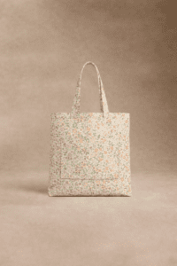 Un sac fourre-tout de forme carrée avec deux anses, présentant un petit motif floral dans des couleurs pastel et une grande poche frontale, est présenté sur un fond neutre.