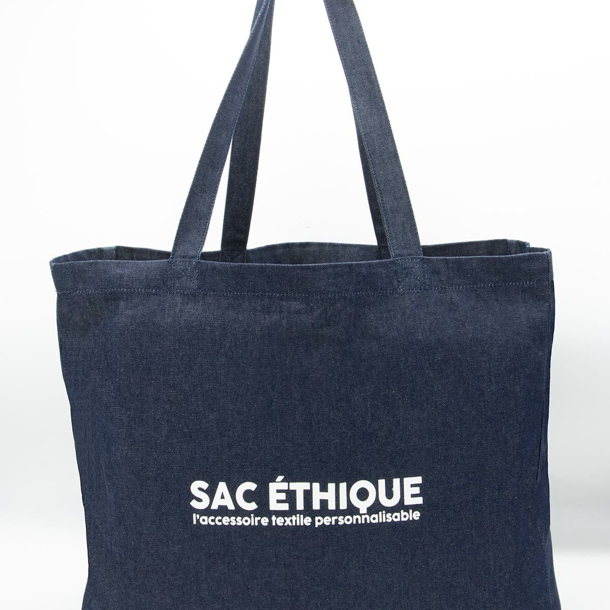 Le Cabas denim est un sac fourre-tout en denim bleu foncé doté de deux anses, sur lequel figure l'inscription « SAC ÉTHIQUE l’accessoire textile personnalisable » en lettres blanches sur le devant. Cet accessoire à la fois élégant et pratique est idéal pour transporter vos affaires quotidiennes et peut être facilement personnalisé selon votre style.