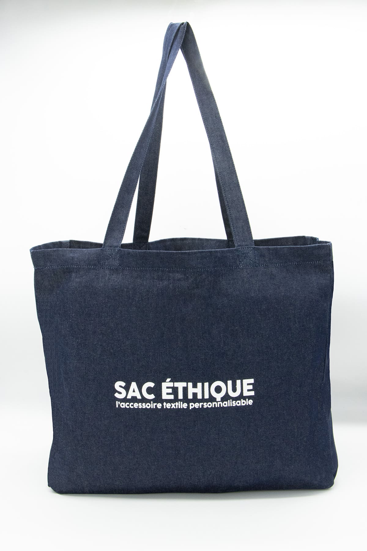 Le Cabas denim est un sac fourre-tout en denim bleu foncé doté de deux anses, sur lequel figure l'inscription « SAC ÉTHIQUE l’accessoire textile personnalisable » en lettres blanches sur le devant. Cet accessoire à la fois élégant et pratique est idéal pour transporter vos affaires quotidiennes et peut être facilement personnalisé selon votre style.